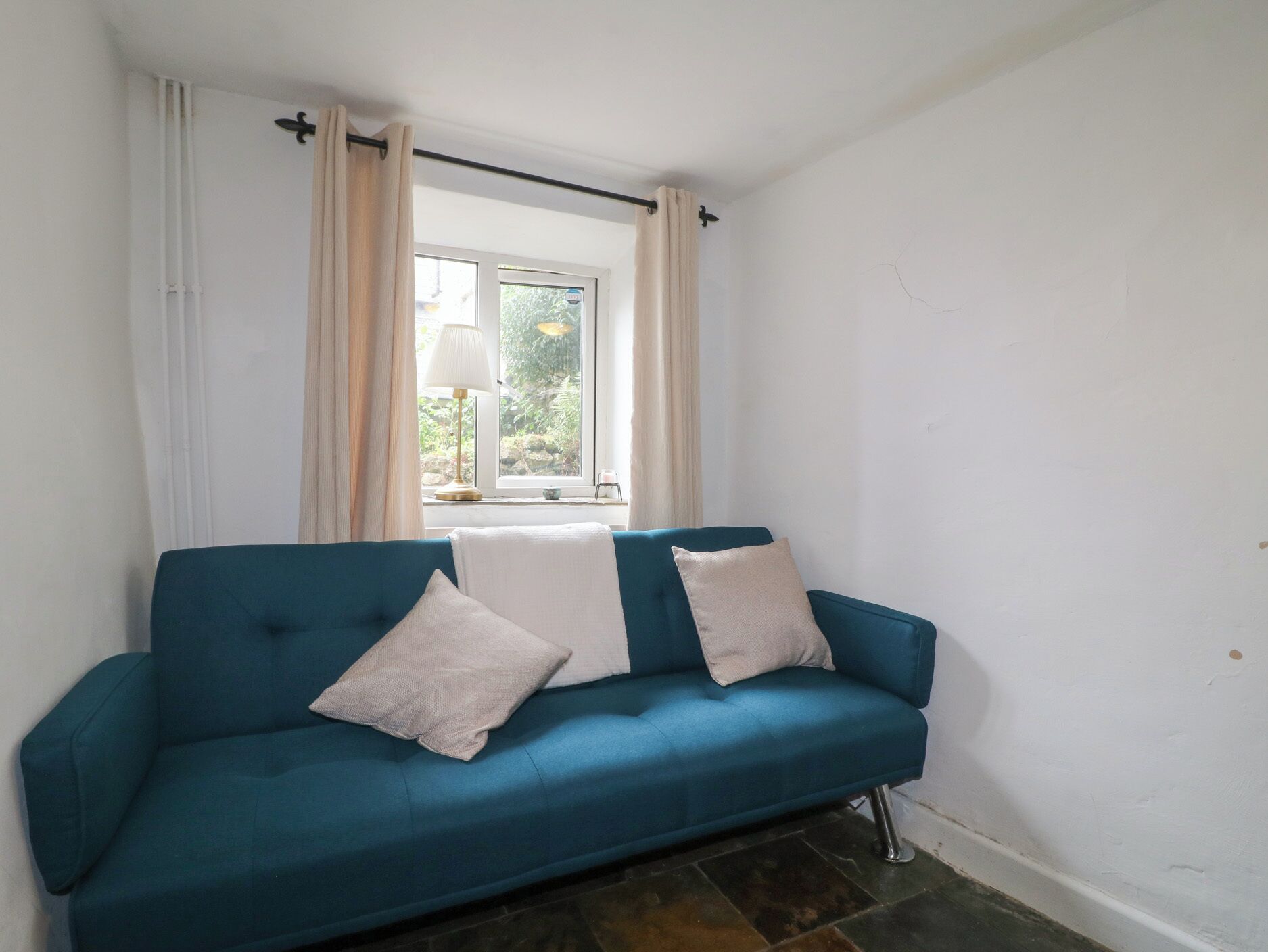 Ein blaugrünes Sofa mit zwei Kissen, ein Fenster mit Vorhängen und eine Lampe auf einem Tisch.