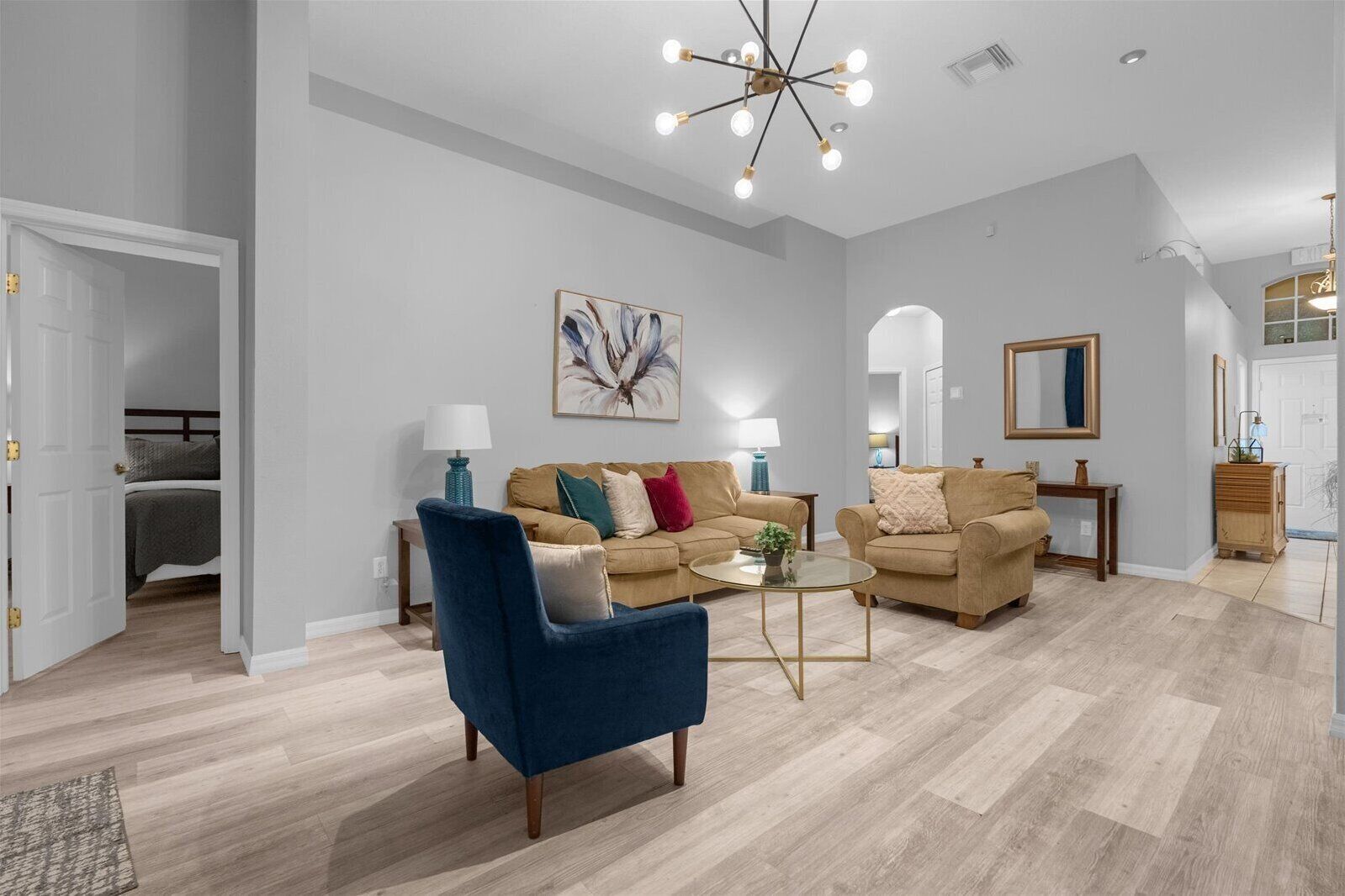 A spacious living room with two sofas, a coffee table, and a blue armchair. There is a painting on the wall and a chandelier above.