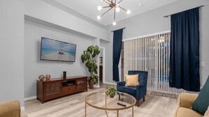 A modern living room with a wooden entertainment center, a blue armchair, a round coffee table, and a large TV on the wall. - Davenport Luxury 8-Guest Villa (Davenport)