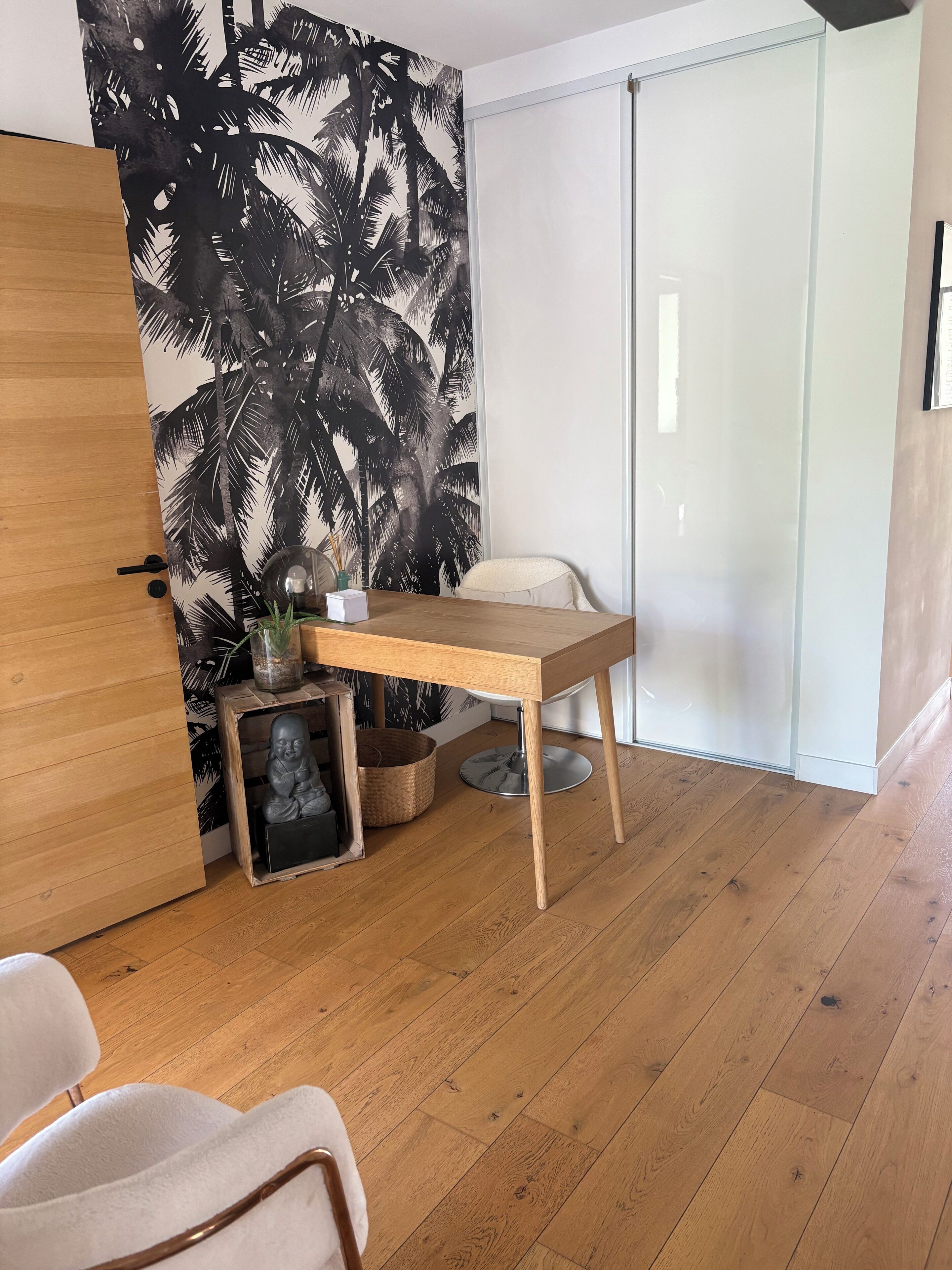 Una habitación con suelo de madera, un escritorio con lámpara, una silla, una pequeña estatua y una pared con estampado de palmeras.