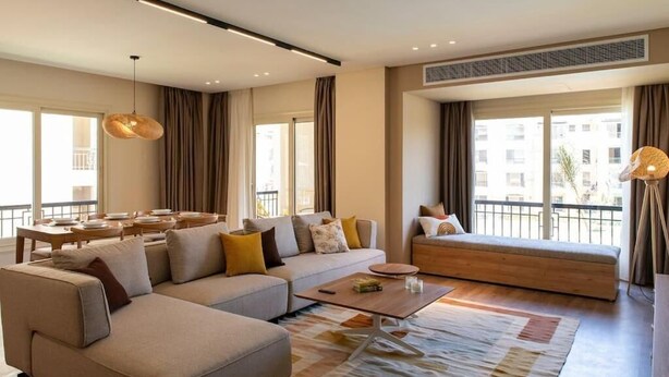 A modern living room with a sectional sofa, a coffee table, and a dining area with a table and chairs. - Kennah Milena | Stone Residence, New Cairo - Families Only (New Cairo)