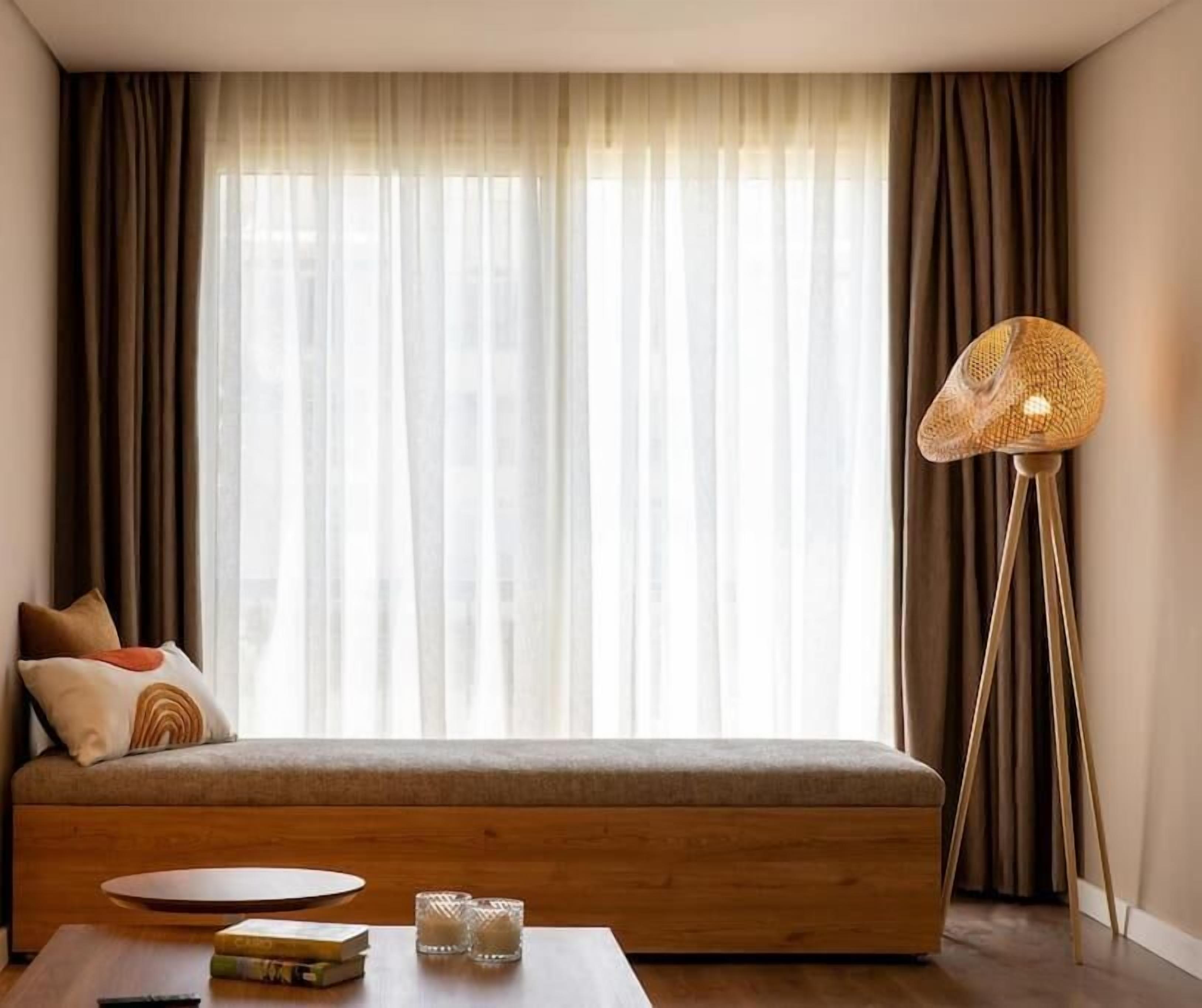 A modern living room with a sofa, a coffee table with books and glasses, a floor lamp, and sheer curtains.