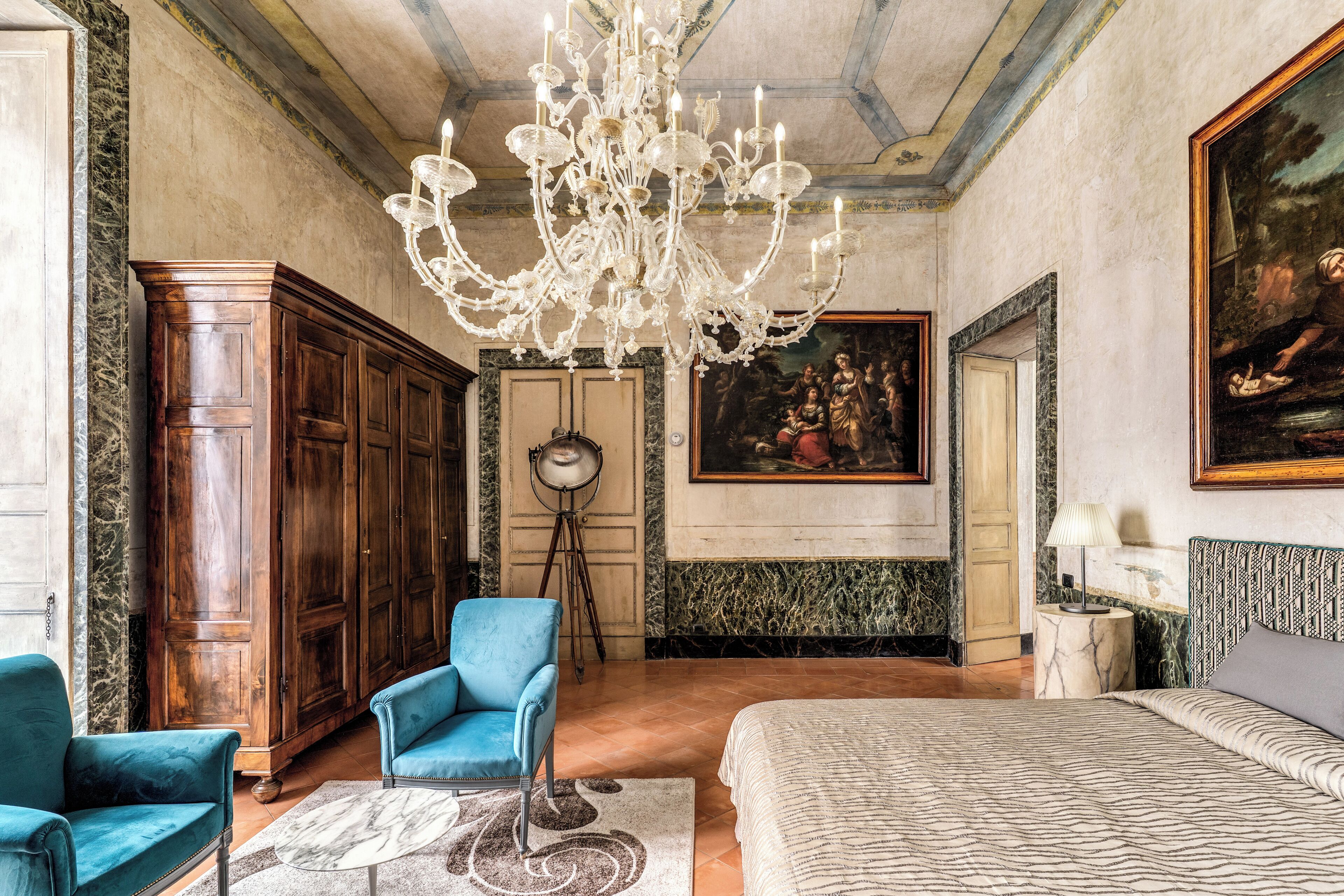 A spacious bedroom with a large bed, a chandelier, a blue armchair, a wooden wardrobe, and a painting on the wall.