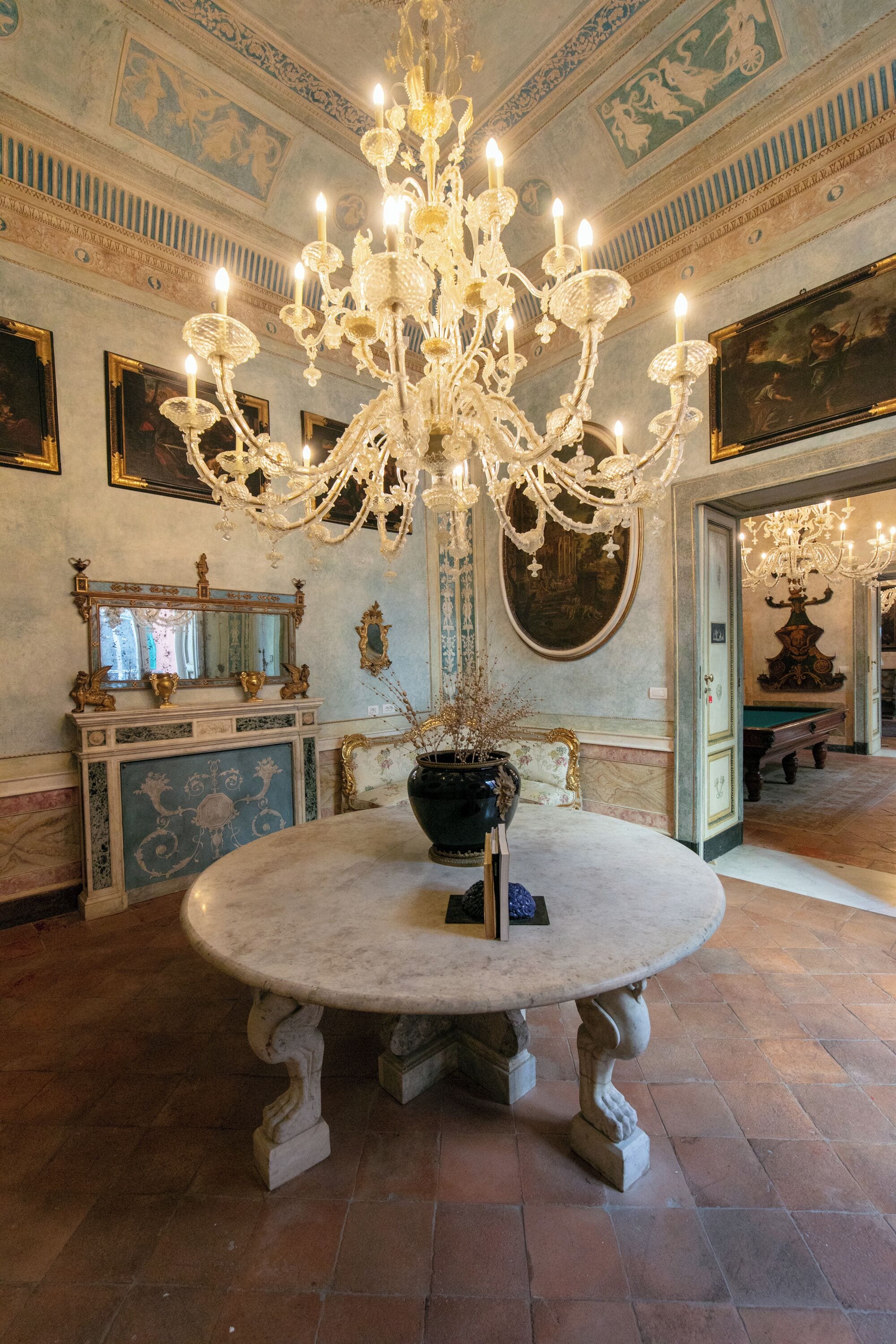 Un'ampia sala con un grande lampadario decorato, un tavolo rotondo in marmo e decorazioni murali in stile classico.
