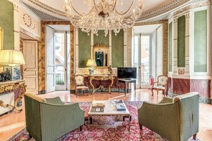 A luxurious living room with a chandelier, green upholstered chairs, a coffee table, and a television.