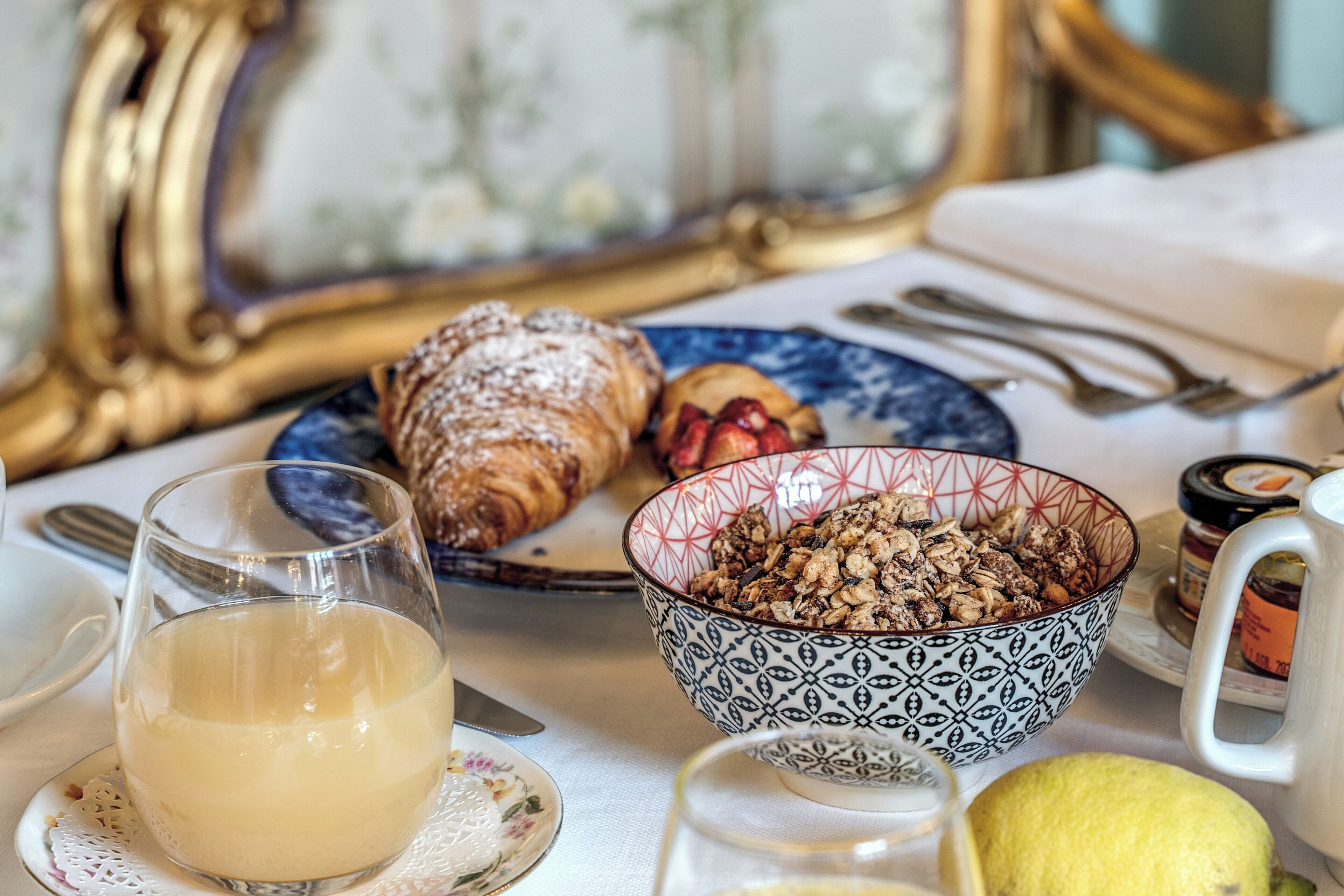 A breakfast setting with a croissant, bowl of granola, a glass of juice, and a lemon on a table.
