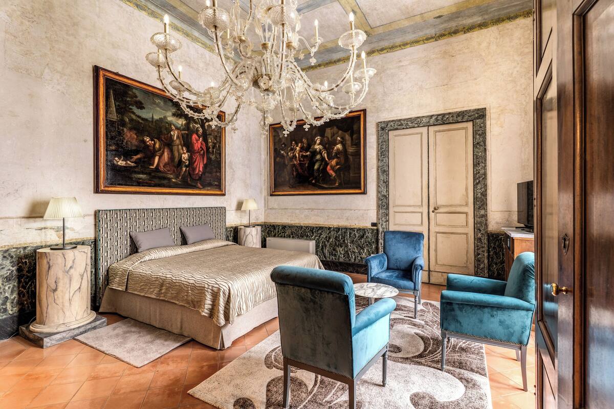 A bedroom with a large bed, two blue armchairs, a chandelier, and two paintings on the wall.