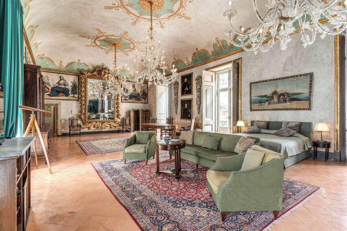 A spacious room with a large bed, green upholstered furniture, a chandelier, and a large painting on the wall.