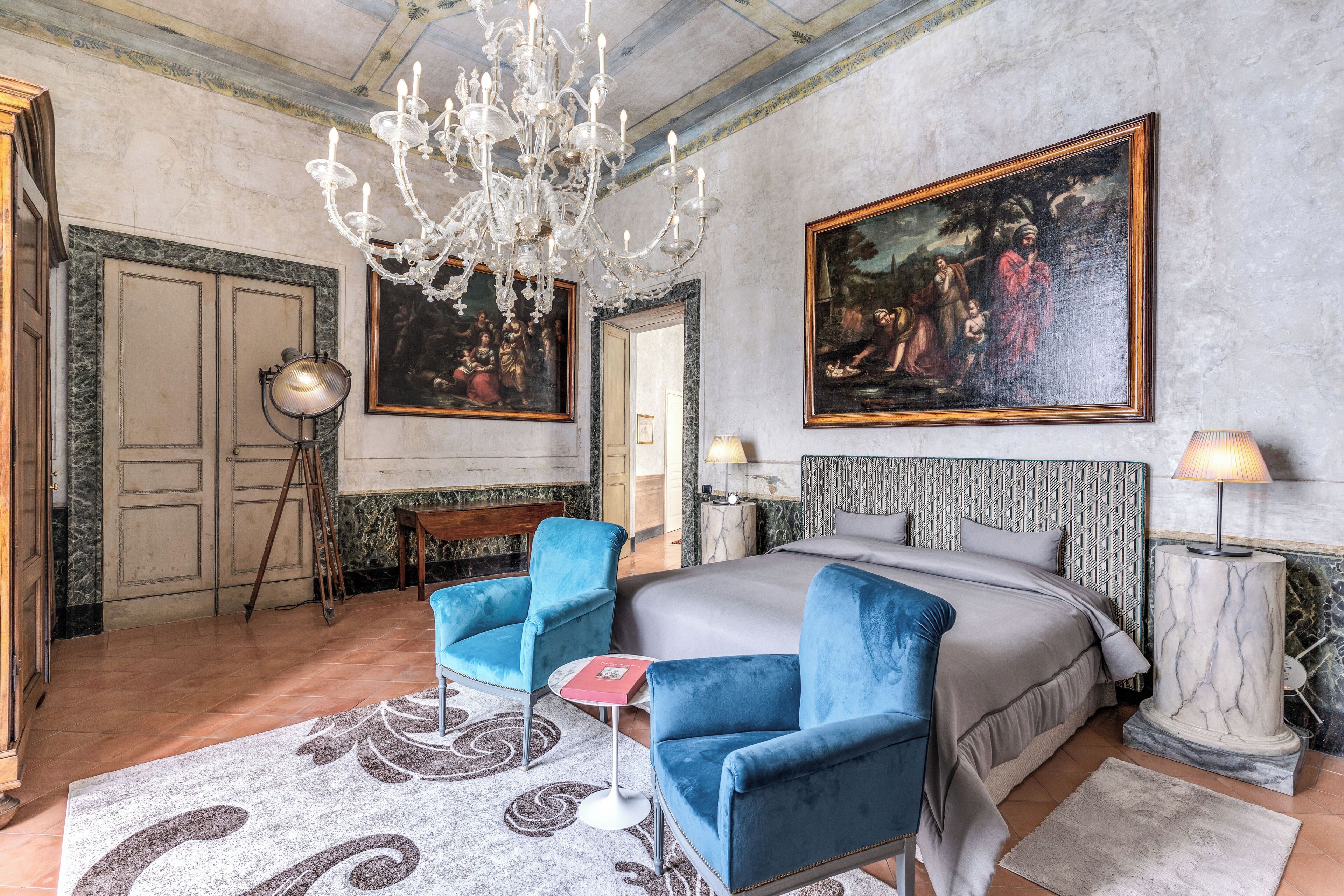 Ein geräumiges Schlafzimmer mit einem großen Bett, zwei blauen Sesseln, einer Chandelüster und klassischen Gemälden an den Wänden.