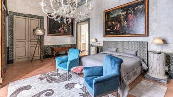 Ein geräumiges Schlafzimmer mit einem großen Bett, zwei blauen Sesseln, einer Chandelüster und klassischen Gemälden an den Wänden.