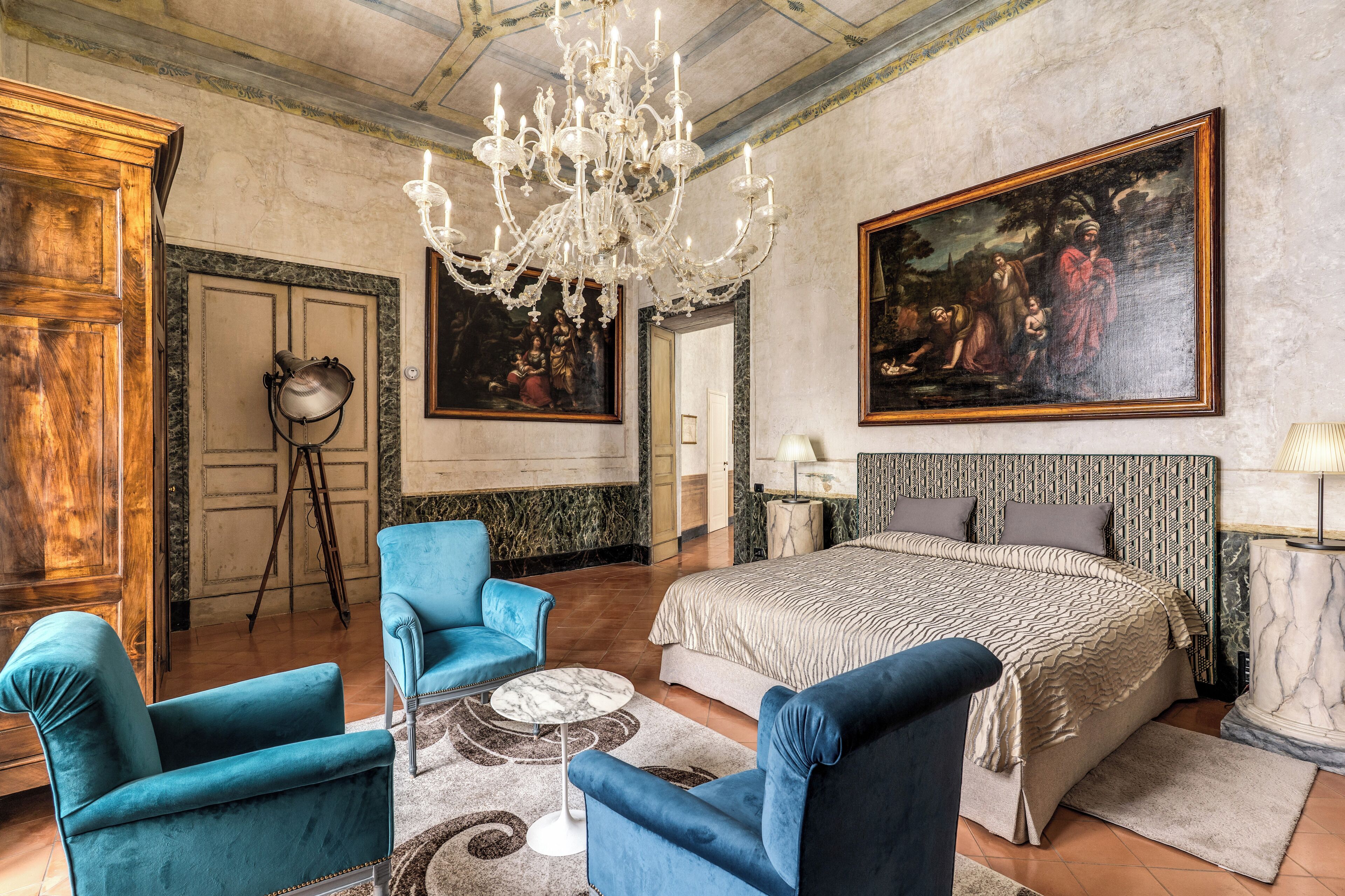 A spacious bedroom with a large bed, a chandelier, and two blue armchairs.