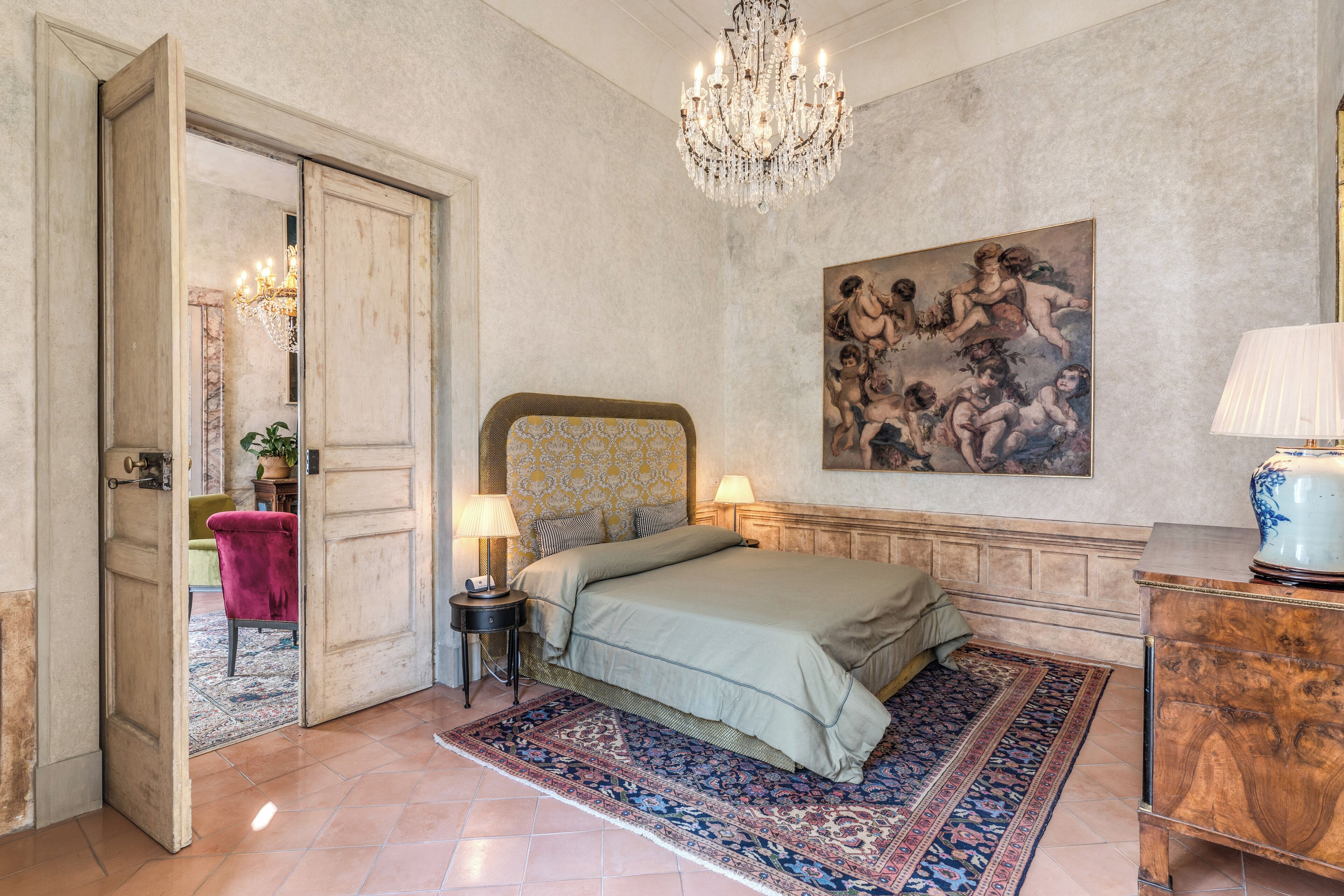 A bedroom with a large bed, a chandelier, a painting, and a nightstand.