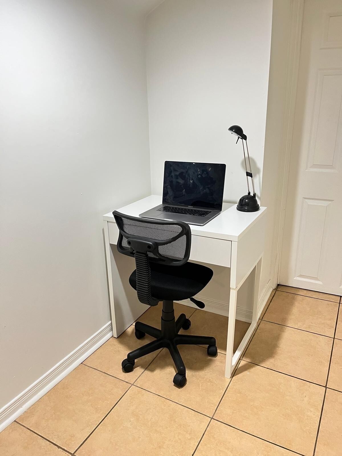 A white desk with a laptop and a desk lamp in a corner room with tiled flooring.