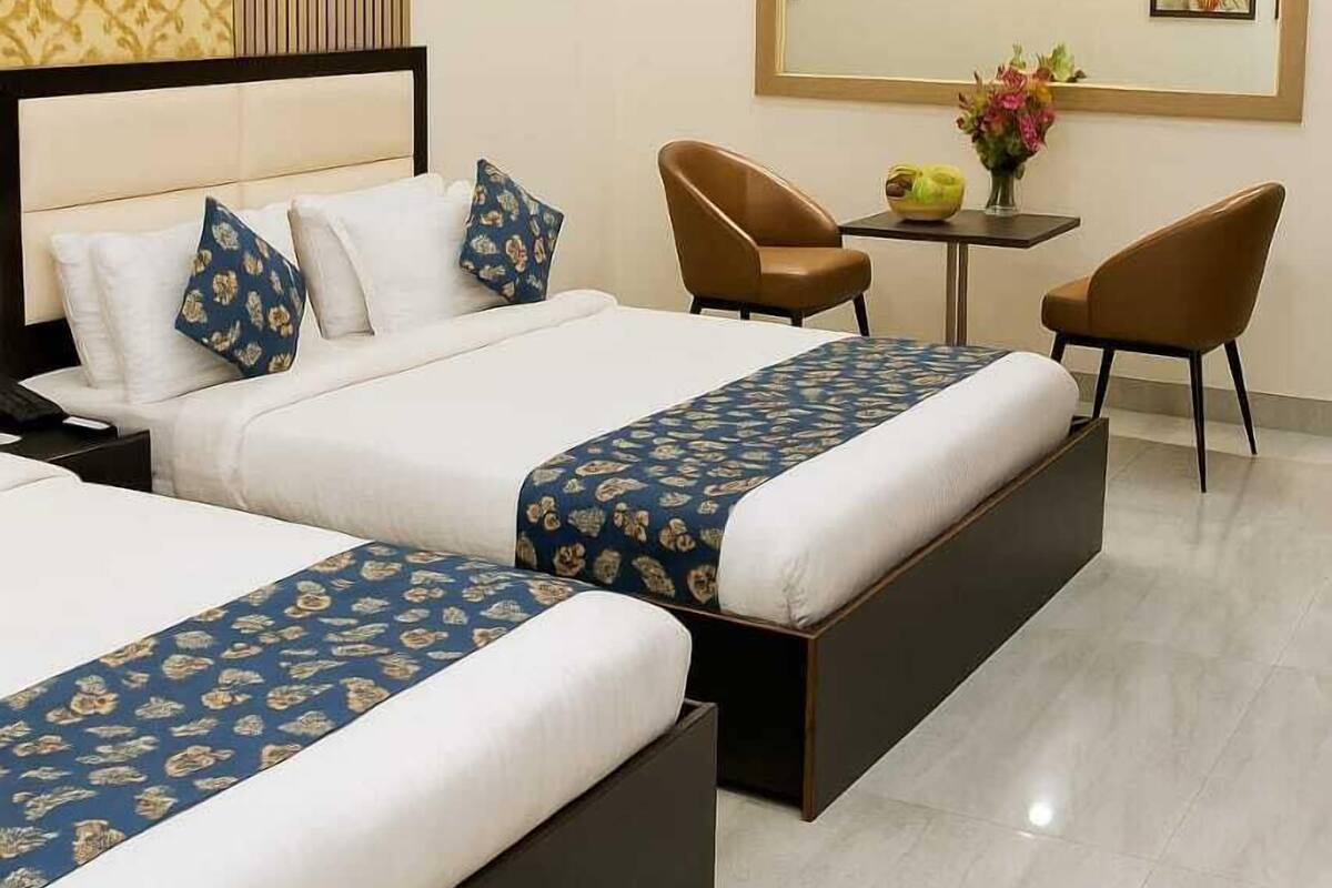 A hotel room with two beds, a desk, chairs, an air conditioning unit, and artwork on the walls.