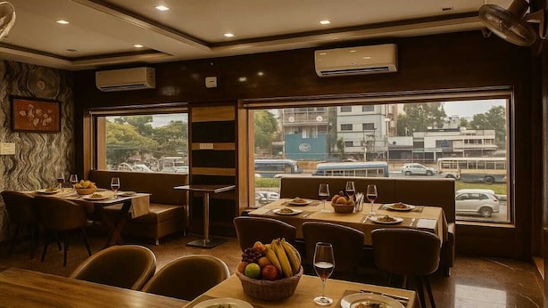A restaurant with tables and chairs, a large window with a view of buildings and vehicles, and air conditioning units on the ceiling. - Bhraj Inn Hotel Udaipur (Udaipur)
