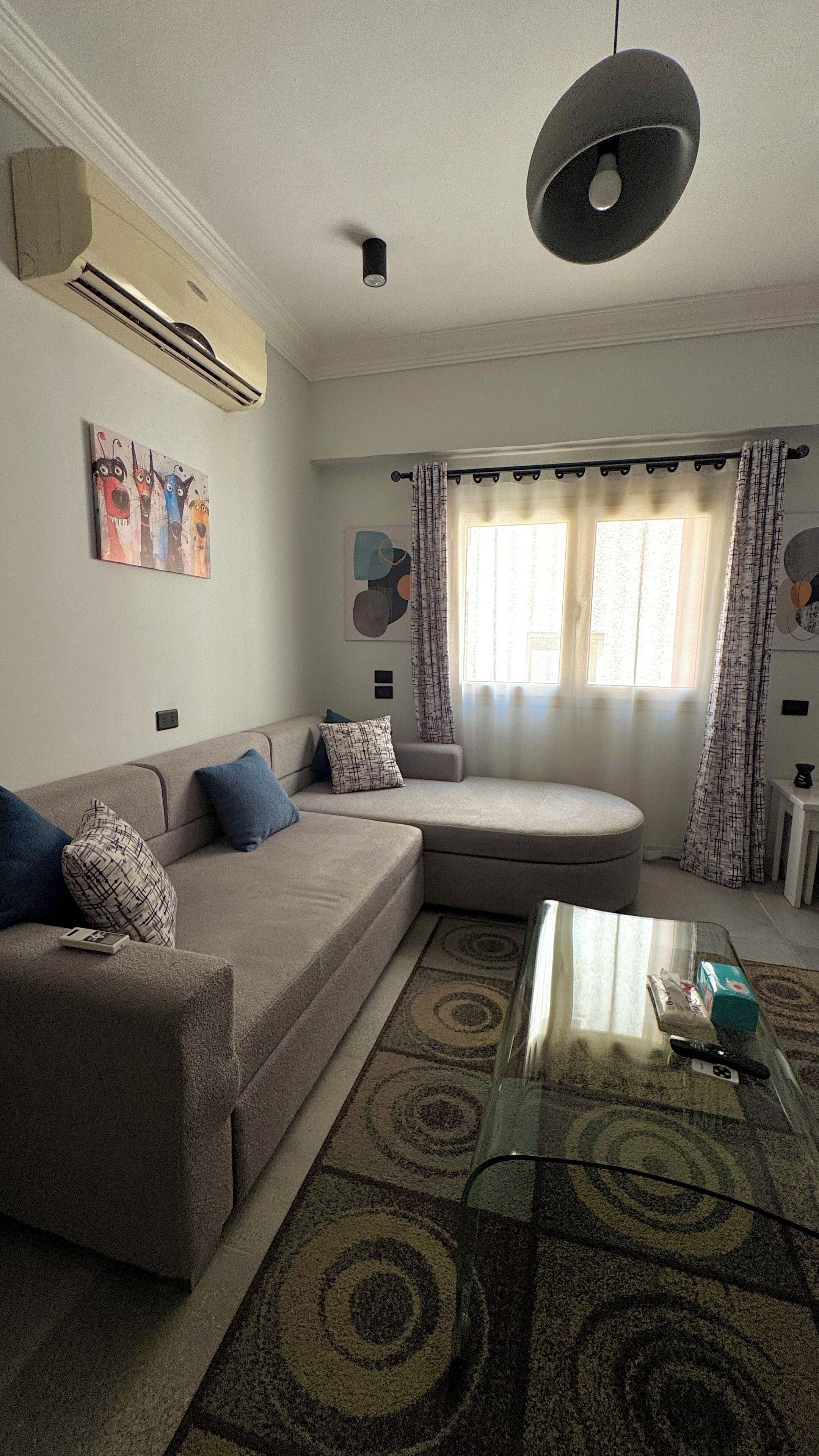 A living room with a sectional sofa, a glass coffee table, and a large window with curtains.