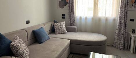 A living room with a sectional sofa, a glass coffee table, and a large window with curtains.
