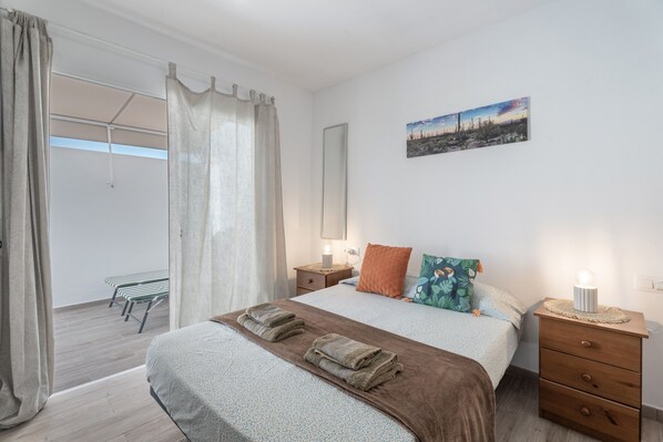A bedroom with a bed, a bedside table with a lamp, a window with curtains, a bench outside, and a painting on the wall.