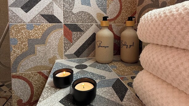 Un baño con baldosas estampadas, dos velas y botellas etiquetadas como 'Shampoo' y 'Body gel'.