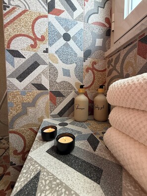 A bathroom with patterned tiles, two candles, and bottles labeled 'Shampoo' and 'Body gel'.