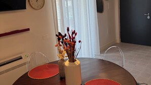 Un comedor moderno con una mesa redonda de madera, dos individuales rojos, un jarrón blanco con flores secas y un reloj fijado a la pared.