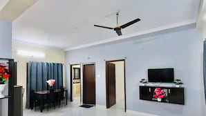 A spacious living area with a ceiling fan, a dining table with chairs, a TV on a black stand, and a door leading to another room.
