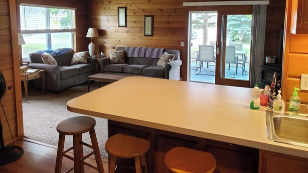 A cabin interior with a wooden interior, a dining area with a table and chairs, a sofa set, and a kitchen area with a sink and bar stools. - Fabulous 2-bedroom Otsego lake cottage in charming Gaylord (Gaylord)