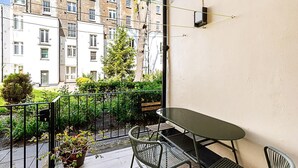 Un balcón con mesa y sillas, una planta en maceta y vista a un edificio con varias ventanas.