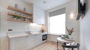 A modern kitchen with white cabinetry, wooden shelves, a built-in oven, and a dining area with a round table and chairs. - The Mezzanine Studio at Bramham (London)