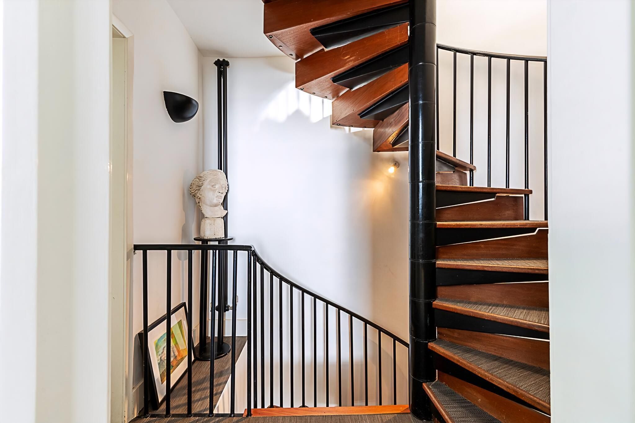 Eine Wendeltreppe mit Holzboden und schwarzen Geländern, eine weiße Wand sowie eine Büstenplastik.