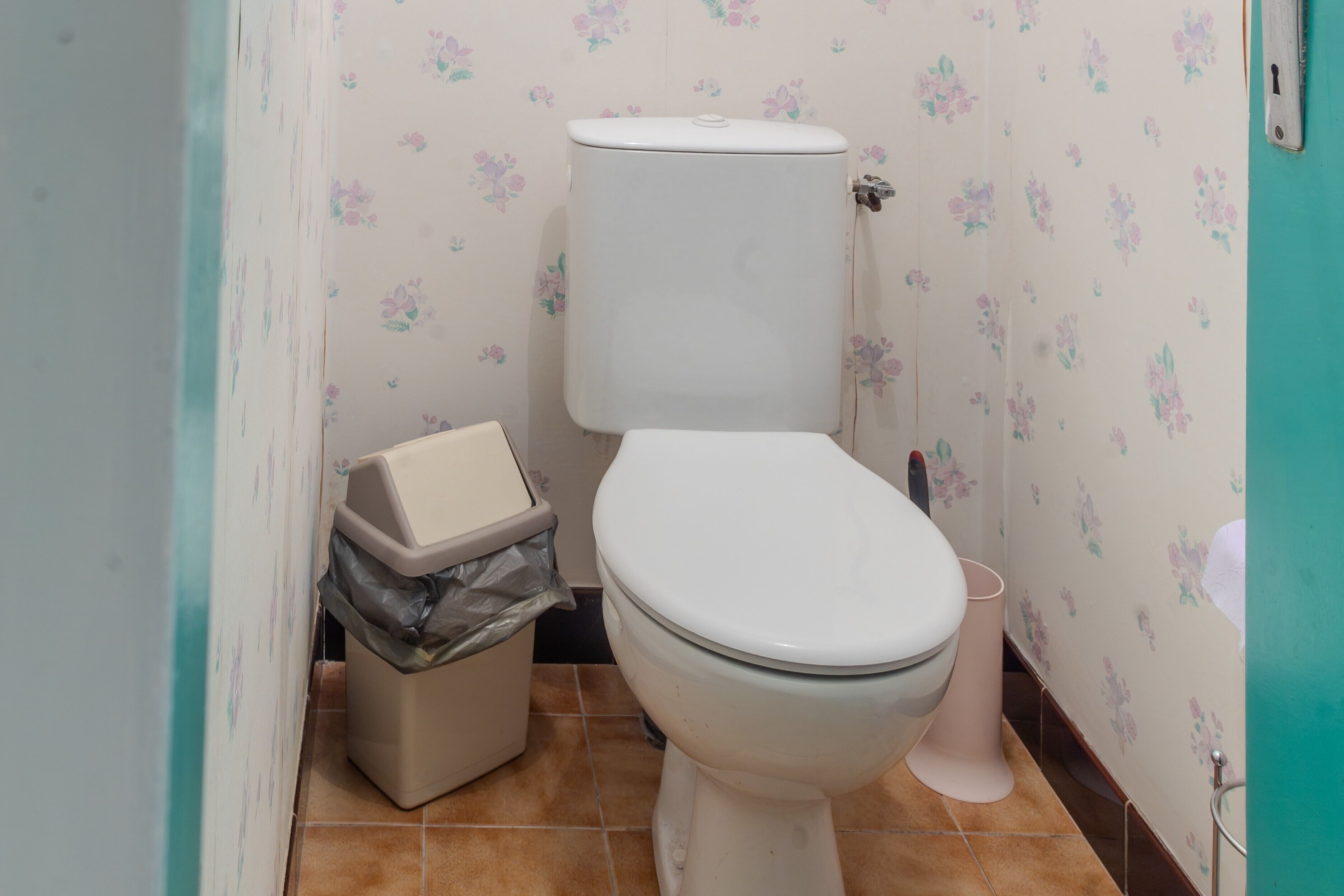 Un baño con inodoro, un bote de basura y una pared con papel tapiz floral.