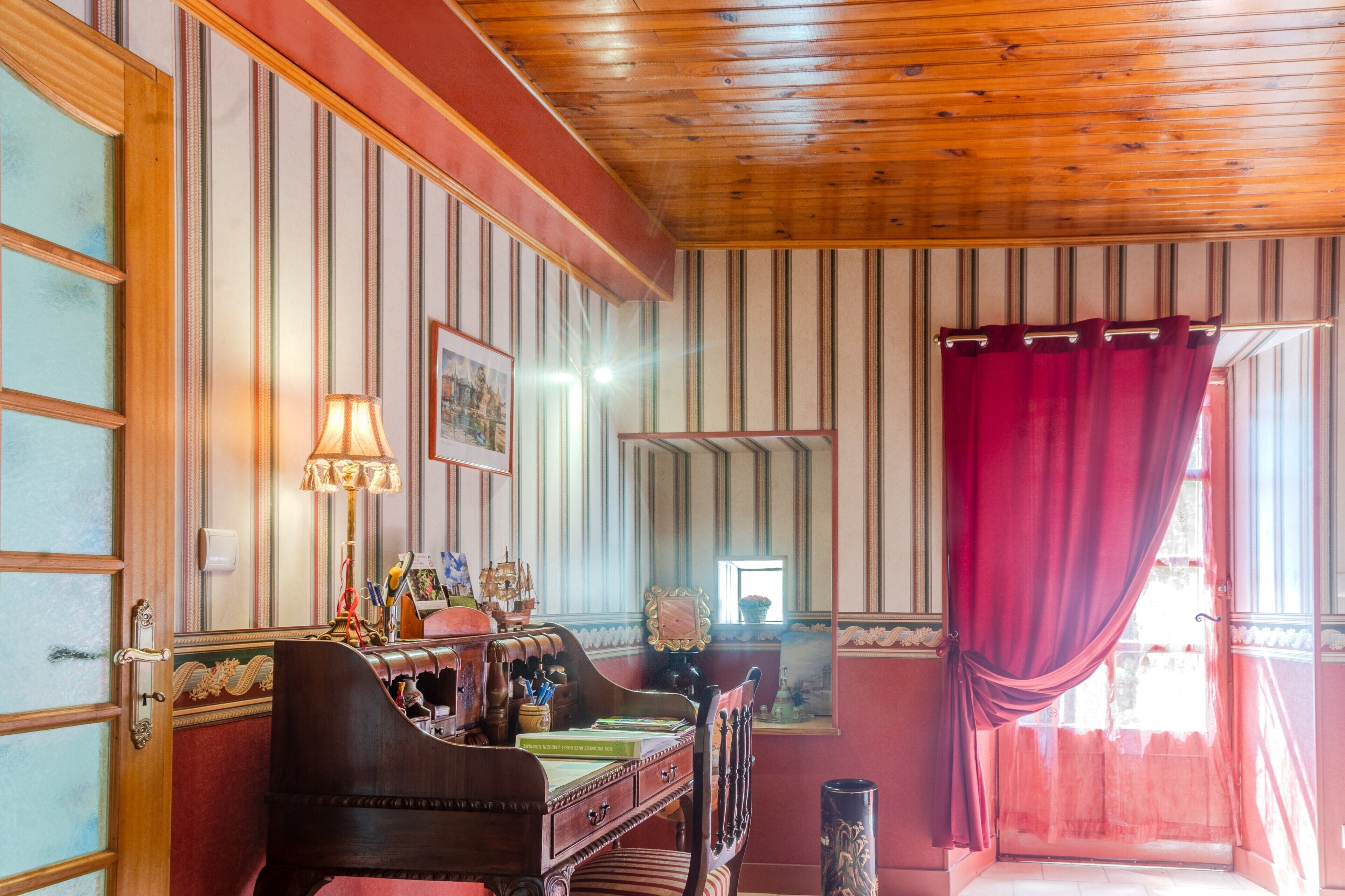 Een kamer met houten lambrisering, een bureau met boeken, een lamp en een raam met een rode gordijn.