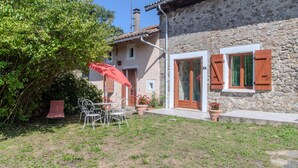 A stone building with a red door, white shutters, and a small porch with a table and chairs. - Holiday Home 'Des Tilleuls' with Private Garden (Saint-Mathieu)