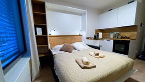 Una camera da letto con un letto, un comodino, una finestra con persiane, una mensola a muro e un angolo cottura.