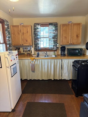 A kitchen with wooden cabinets, a refrigerator, a microwave, a stove, and a window with curtains.
