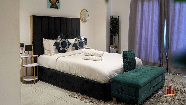 A modern bedroom with a large bed, a bedside table, a mirror, and a green ottoman.