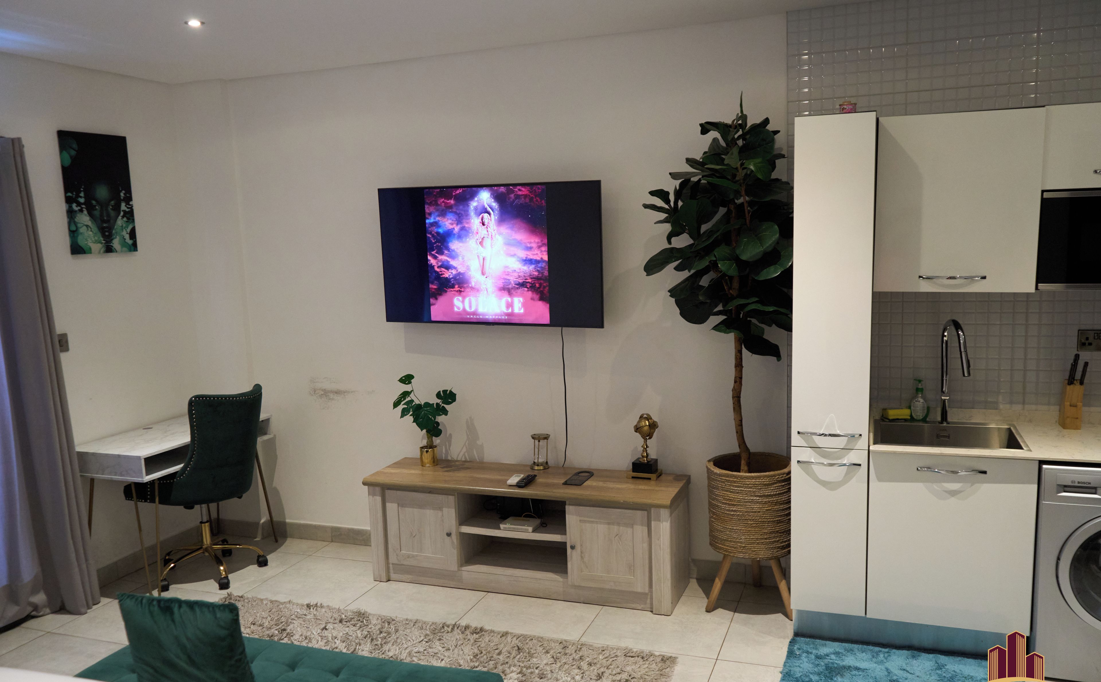 A modern living room with a flat-screen TV, a green sofa, a wooden coffee table, and a kitchen area with a washing machine.