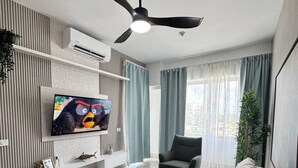 Un salon moderne doté d’un téléviseur à écran plat, d’un ventilateur de plafond, d’un canapé gris, d’une table basse en bois et d’une grande fenêtre avec des rideaux.