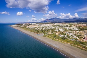 Vista aérea de un pueblo costero con playa, edificios residenciales y una montaña al fondo.