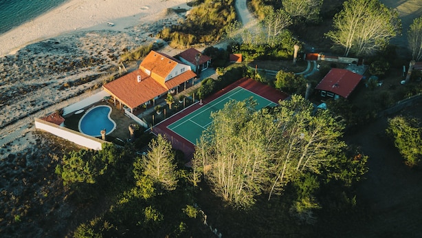 An aerial view of a coastal property with a swimming pool, tennis court, and a house with a red roof.