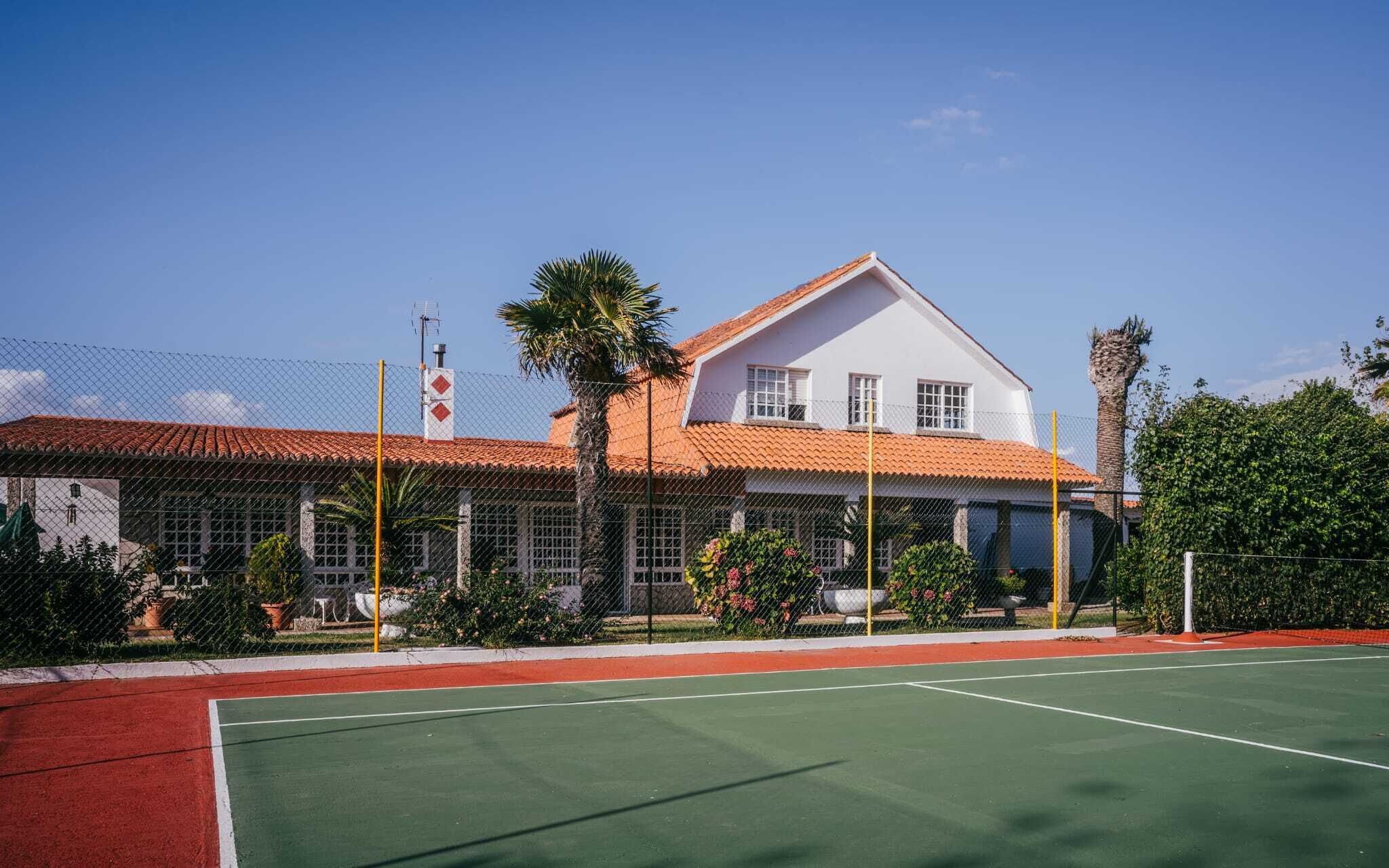 Una cancha de tenis con superficie roja y verde, rodeada por una valla, y un edificio blanco con techo de tejas rojas al fondo.