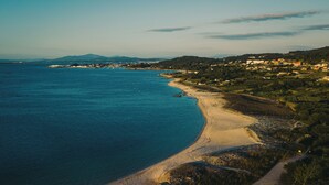 A coastal landscape with a sandy beach, clear blue sea, and a hilly town in the background.