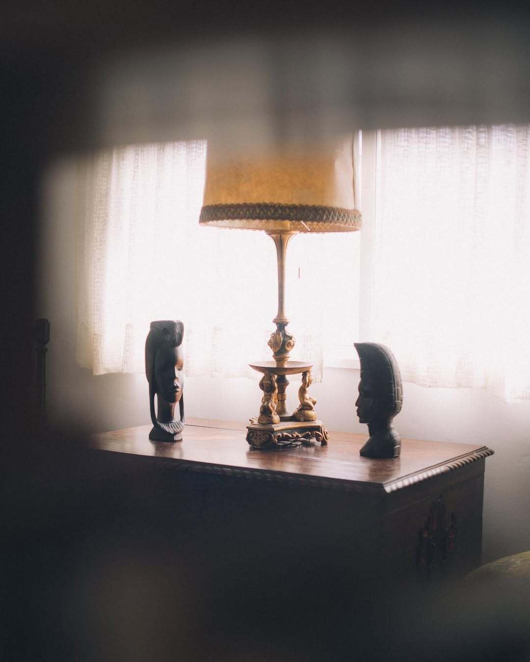 A table with a lamp and two sculptures in a dimly lit room with a window in the background.
