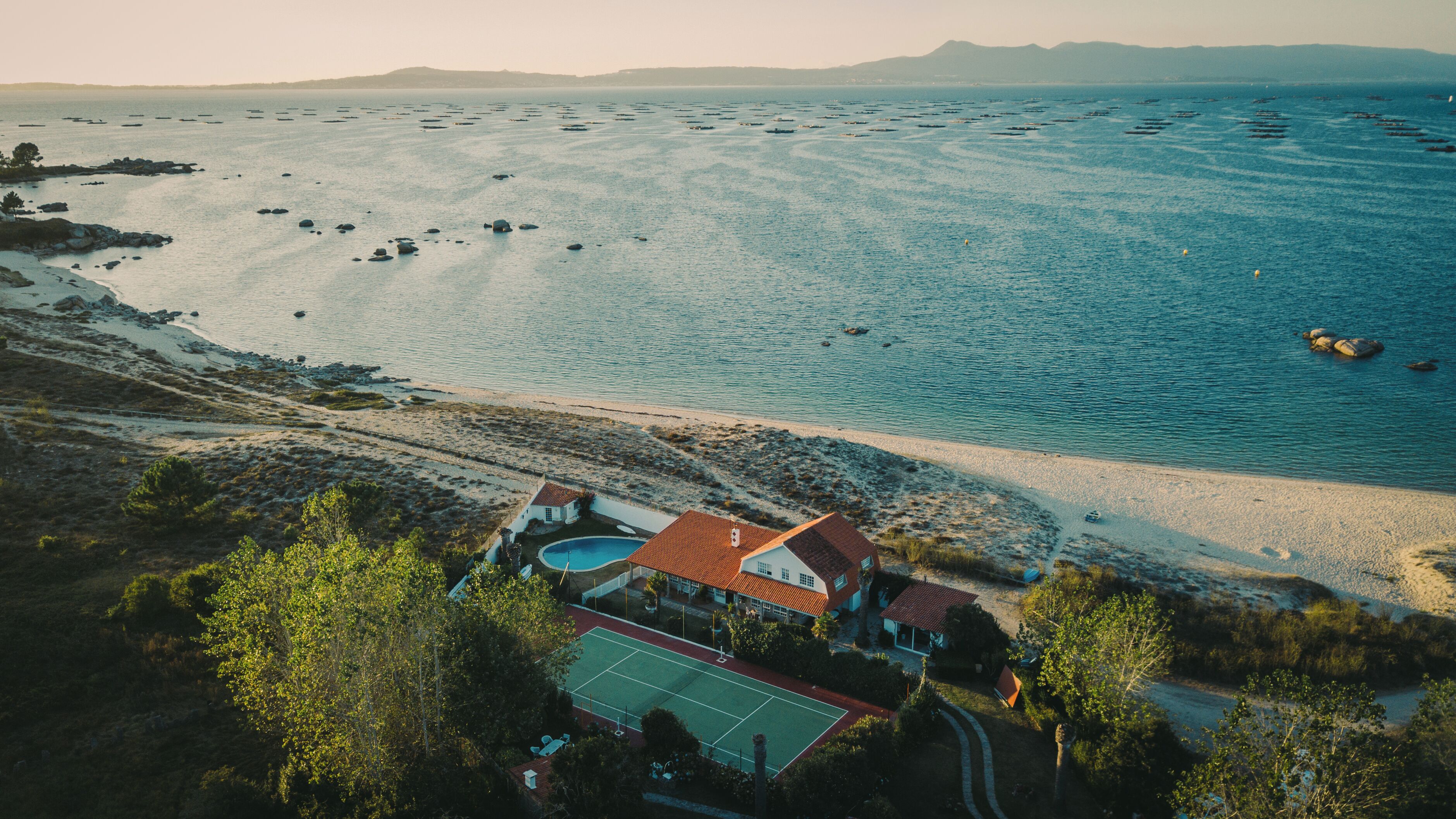 An aerial view of a coastal property with a tennis court, beach access, and a view of the sea.