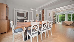 A spacious dining area with a wooden table and white chairs, a kitchen in the background, and large windows offering a view of greenery. - 19 min to the center - 250 m² / 2,691 sq ft - top villa from 1923 - 4BR 3,5BA (Gräfelfing)