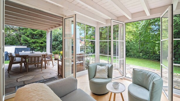 A spacious room with a large sliding glass door leading to an outdoor patio area. Inside, there is a grey sofa, a round armchair, and a small round table. - 19 min to the center - 250 m² / 2,691 sq ft - top villa from 1923 - 4BR 3,5BA (Gräfelfing)