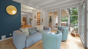 A spacious living room with a large sofa, a coffee table, and a blue ottoman. There is a bookshelf filled with books and a dining area with a table and chairs. - 19 min to the center - 250 m² / 2,691 sq ft - top villa from 1923 - 4BR 3,5BA (Gräfelfing)