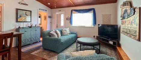 A cozy living room with a wooden ceiling, a green sofa, a dining table with chairs, a television, and a window with blue curtains.