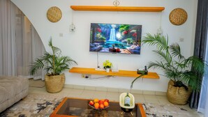 A living room with a large TV mounted on the wall, a wooden shelf below it, and a coffee table with fruit and a bottle. - 2BR Apt, 6-Min Walk to Diani Beach, Kids Playground, Near Ukunda Airport. (Diani Beach)