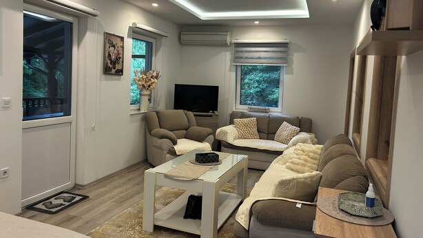A modern living room with a sofa, armchair, and a coffee table. There are two windows with blinds, a flat-screen TV, and a ceiling light fixture. - Terrific villa with AC in charming Sarajevo (Sarajevo)